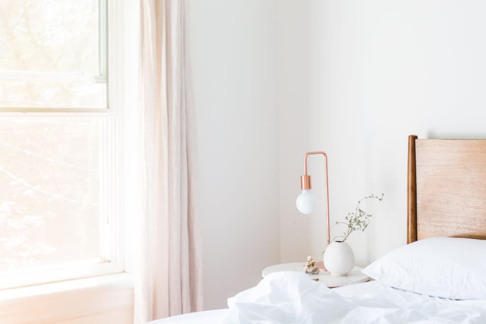 Kamar tidur yang terang dan sepi dengan tirai tipis, lampu meja, dan cahaya pagi dari jendela.